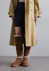 Brown knee-high boots made of suede with a soft fur lining. Paired with a dark denim skirt and a beige trench coat.