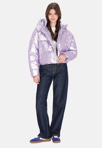 Metallic lilla pufferjakke med hætte, lynlåslukning og quiltet design, kombineret med mørkeblå jeans og blå sneakers.