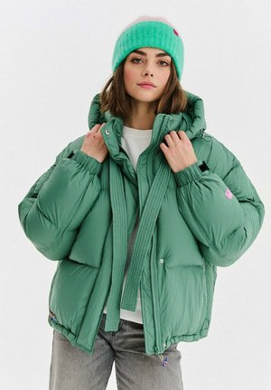 Zielona puchowa kurtka o grubej, pikowanej fakturze, z przednim zamkiem błyskawicznym, regulowanym kapturem i bocznymi kieszeniami. Do tego lekko zielona i różowa dziana czapka.