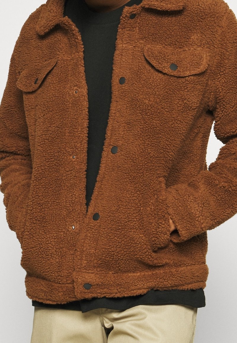 Chaqueta de forro polar marrón con una superficie texturizada, que presenta un cuello con solapa, botones a presión y dos bolsillos en el pecho. Llevada sobre una camiseta negra.