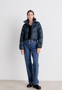 Puffet, kort jakke i mørkeblå nylon med høy krage. Kombinert med høy midje blå jeans og svarte platåsko. Minimalistisk design.