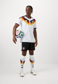 Biely futbalový dres s červenými, čiernymi a žltými akcentmi, čierne šortky a ponožky po kolená s živým vzorom. Drží futbalovú loptu.