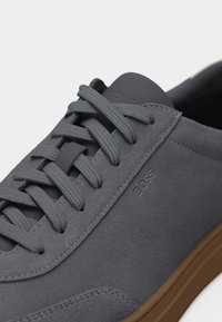 Gros plan sur une basket en daim gris avec des lacets assortis et une semelle marron, ornée du logo BOSS embossé sur le côté.