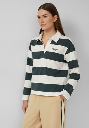 Frau trägt ein langärmliges, weißes und dunkelgrünes gestreiftes Rugby-Shirt mit dem Text "Fullness" und beige Hosen mit schwarzen und weißen Seitenstreifen.