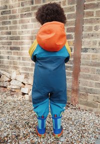 Kleurrijk regenpak met oranje capuchon, blauwe en gele schouders, en blauwe broekspijpen. Inclusief waterdichte laarzen met kleurrijke accenten en patronen.