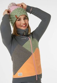 Giacca in pile con un design asimmetrico che presenta pannelli arancioni, verdi e grigi; dettagli in rosa sulla cerniera e sul logo; indossata con un berretto a maglia.