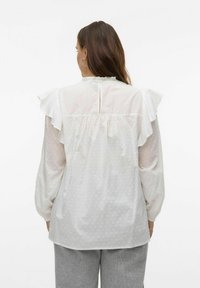 Blouse blanche texturée avec des épaules volantées, un dos plissé et une fermeture en forme de clé. Fabriquée à partir d'un matériau léger et respirant.