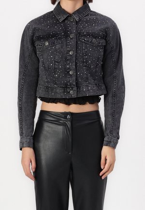 Femme portant une veste en jean noire courte avec des clous argentés, des fermetures à boutons, et un pantalon en cuir noir taille haute sur fond blanc.