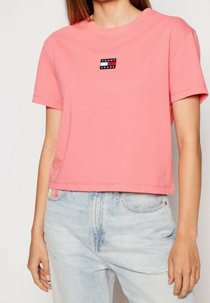 Kobieta nosząca różowy krótki krótki t-shirt z małym czarno-białym logo Tommy Jeans na piersi, w połączeniu z jasnoniebieskimi dżinsami.