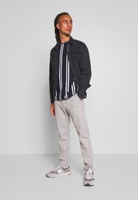 Chaqueta de mezclilla negra sobre una camiseta de rayas azul marino, pantalones de chándal grises y zapatillas grises con detalles en blanco. El modelo está de pie contra un fondo liso.
