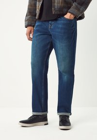 Tmavomodré rifle z denimu s rovnými nohavicami, piatimi vreckami a jemným vyblednutím. Nosené s čiernymi šnurovacími topánkami a vyhrnutými lemami.