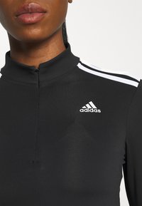 Zbliżenie na osobę w czarnej rozpinanej bluzie Adidas z białym logo i trzema białymi paskami na ramieniu.