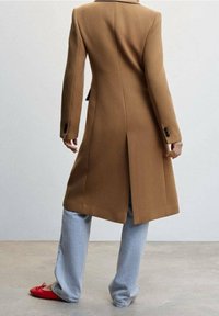 Cappotto lungo marrone con una vestibilità sartoriale, caratterizzato da uno spacco sul retro, abbinato a jeans a gamba larga blu chiaro e ballerine rosse.