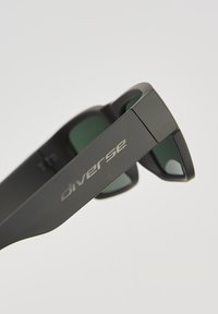 Okulary przeciwsłoneczne z matową czarną oprawą, ciemnozielonymi soczewkami i srebrnym logo "diverse" z boku. Elegancki, nowoczesny design o minimalistycznym profilu.