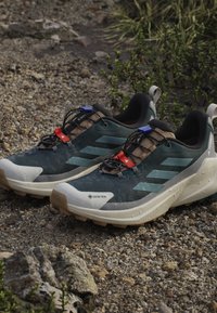 Pair of dark green and beige Gore-Tex trail running shoes with laces and pull tabs on rocky outdoor ground.