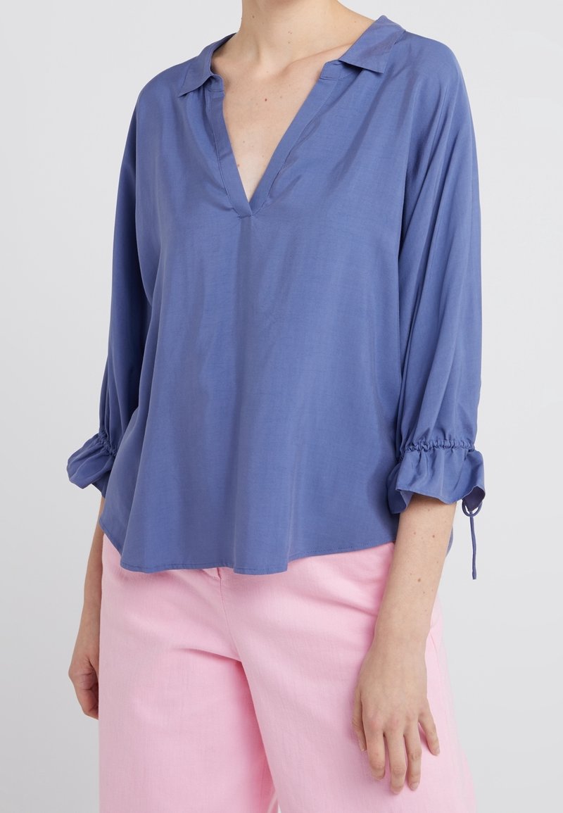 Blusa blu chiaro, dalla vestibilità ampia, con scollatura a V e maniche a tre quarti. Tessuto morbido con polsini arricciati. Indossata con pantaloni rosa pallido.