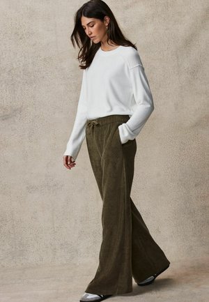Pulover alb cu mânecă lungă și pantaloni cu talie înaltă din catifea în nuanță de verde măsliniu, cu șnur. Textură moale și croială lejeră, asortați cu pantofi negri și gri.