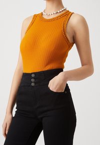 Orange ribbad ärmlös topp med spetsdetaljer vid axlarna, kombinerad med högmidjade svarta byxor med knappstängning framtill.