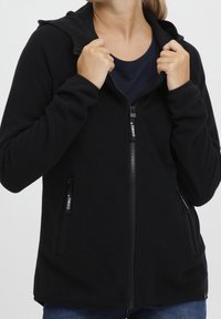 Haut en polaire noir zippé avec une surface texturée, capuche ajustable et deux poches latérales. Présente un logo sur les zip et une coupe décontractée.