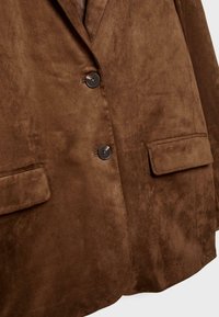 Blazer en suède marron avec deux poches avant, fermeture à deux boutons et texture lisse, présentant une coupe ajustée et un design à revers classique.