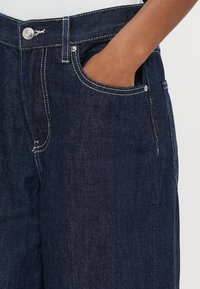 Tumši zili džinsa džinsi ar kontrastējošām šuvēm, monētu kabatu un standarta jostasvietu ar priekšējo aizdari.