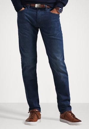 Jeans de mezclilla azul oscuro con un diseño de corte recto, cinco bolsillos y un cinturón de cuero marrón. Combinados con zapatillas de cordones marrones.