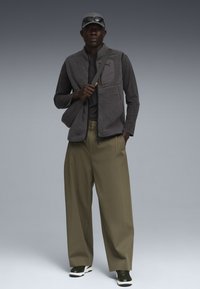 Fleece-Weste in Dunkelgrau mit einer Reißverschluss-Tasche, über einem schwarzen Oberteil getragen. Olivgrüne, weit geschnittene Hosen und dunkle Sneaker mit weißen Akzenten.