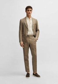 Lys brun linjakke med slått lapel, tilpassede bukser og en kremfarget skjorte med knapper. Brune slip-on sko fullfører antrekket.
