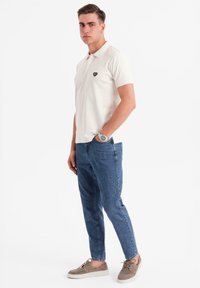 Vit polo tröja med en svart logotyp, matchad med ljusblå avsmalnande jeans och gråcasual skor. Accessoarer inkluderar en silverfärgad klocka.