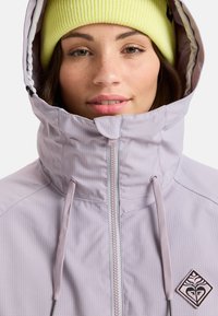 Lichtpaarse jas met een hoge kraag, ritsluiting, trekkoorden en een driehoekig logo. Gekocht met een felgele geribde beanie.