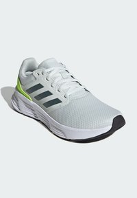 B baskets de sport gris clair avec une tige texturée, une semelle blanche, un embout en caoutchouc noir et trois rayures noires sur le côté, accentuées par un détail vert lime.