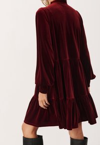Burgundy sametti mekko, jossa on pitkät, puffatut hihat, pitsireunus kauluksessa ja kerroksellinen helma. Kankaan pinta on pehmeä ja sileä.