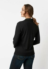 Schwarze, gerippte Jacke mit tailliertem Schnitt, die einen Notchkragen und lange Ärmel aufweist. Über dunkler Jeans getragen, zeigt sie ein elegantes Design.