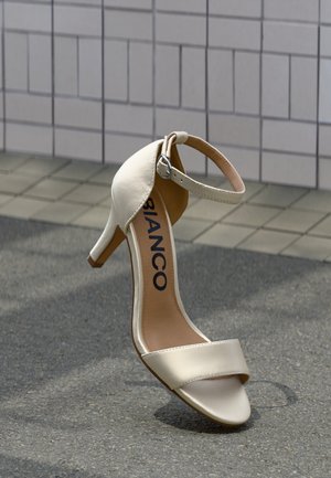 Beige højhælet sandal lavet af glat materiale, med en ankelsnor med sølvspænde og tre smalle frontremme.