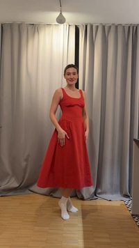 Femme portant une robe midi rouge sans manches et des chaussettes blanches, debout sur un sol en bois devant des rideaux gris à l'intérieur.