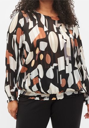 Femme portant un chemisier à manches longues avec des formes géométriques abstraites noires, blanches, marron et beige associé à un pantalon noir.