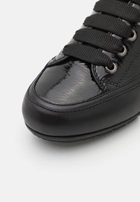 Gros plan sur l'avant d'une basket noire en cuir verni brillant et sections mates lisses, avec des lacets noirs à travers des œillets argentés.