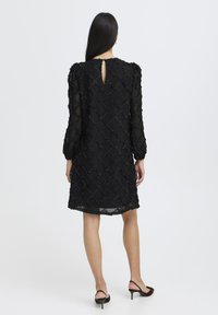 Sorte blondekjole med lange ærmer, der har et diamantmønster, detaljer med nøglehul bagpå og en A-formet silhuet. Accentueret med teksturerede udsmykninger.