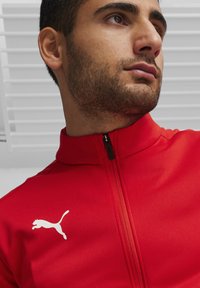Rote Zip-Jacke aus glattem Stoff mit einem weißen Puma-Logo auf der linken Brust und einem hohen Kragen für zusätzlichen Schutz.