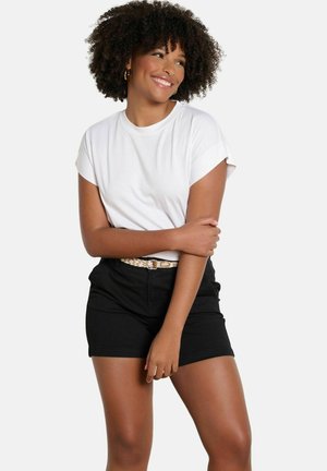 Lächelnde Frau mit lockigem Haar trägt weißes kurzärmeliges Hemd und schwarze Shorts mit geflochtenem Gürtel, steht mit verschränkten Armen vor einem einfarbigen Hintergrund.