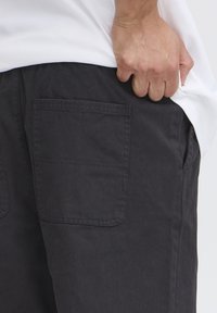 Dark gray casual pants with a single back pocket and subtle stitching details, paired with a white shirt, featuring a hand resting on the side.