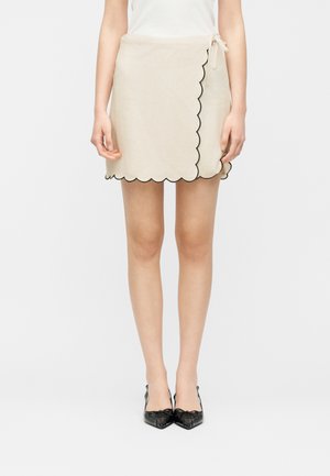 Femme portant une jupe portefeuille beige à bords festonnés avec une bordure noire et des chaussures slingback à bout pointu noires, debout devant un fond blanc.