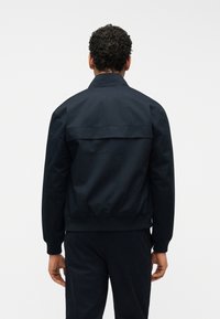 Άνδρας με σγουρά μαλλιά, ντυμένος με μαύρο μπουφάν και μαύρο παντελόνι, στέκεται γυρισμένος με την πλάτη σε απλό λευκό φόντο.