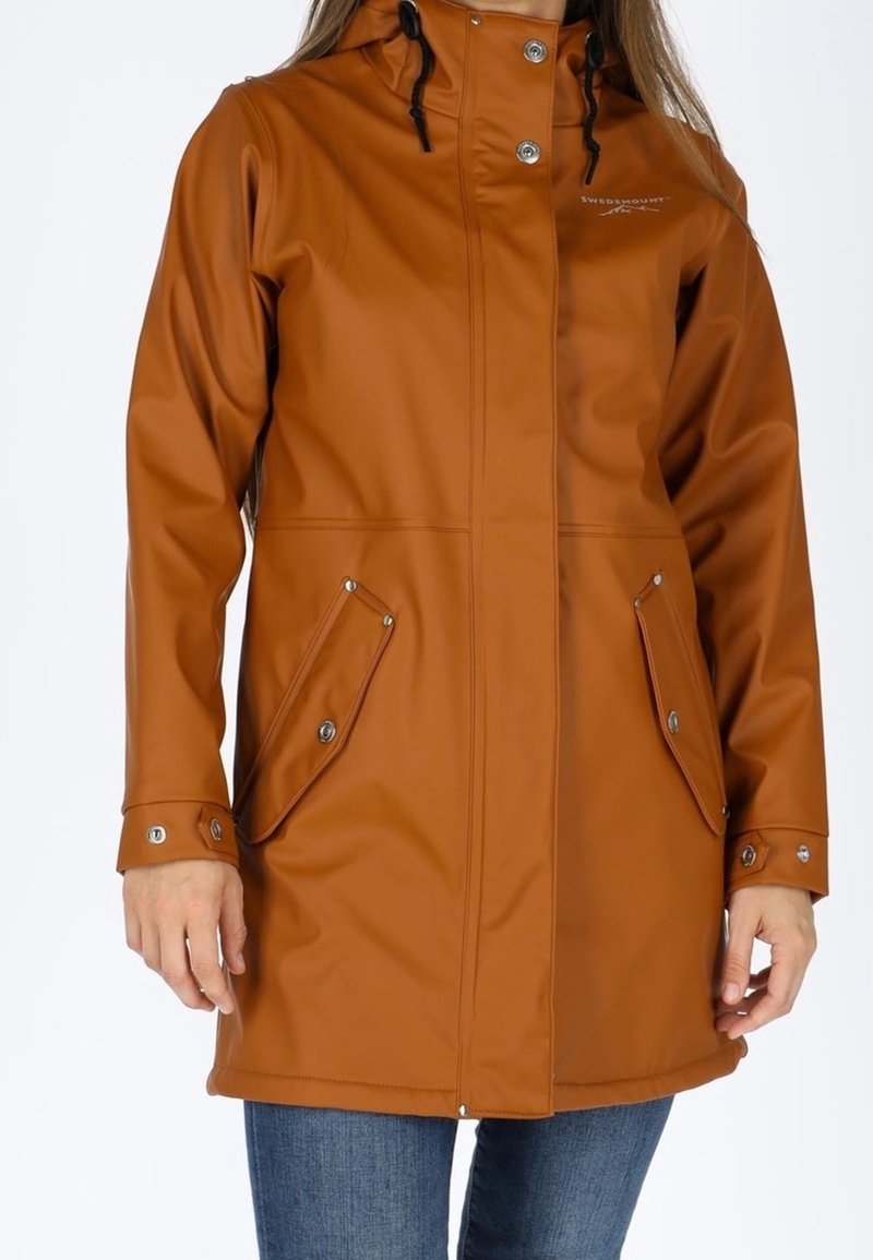 Femme portant un imperméable marron mi-long avec des pressions à l'avant, des poches à rabat et une capuche, assorti à un jean bleu.