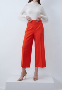 Haut blanc à manches longues associé à un pantalon orange taille haute à jambes larges. Le mannequin est debout sur un piédestal blanc, portant des talons beiges.