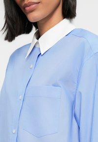 Ανοιχτό μπλε πουκάμισο με λευκό γιακά, με τσέπη στο στήθος και απαλή υφή, φωτογραφημένο από τον ώμο και πάνω.