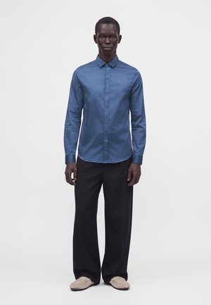 Mandlig model står med front mod kamera iført en blå skjorte med knapper, sorte bukser med vide ben og beige slip-on sko på en ensfarvet hvid baggrund.