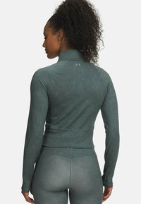 Woman with curly hair wearing a fitted dark green long-sleeve athletic top and matching leggings, viewed from the back.