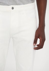 White denim jeans with a slim fit, featuring a silver button closure and five-pocket design. Textured fabric, no visible patterns.