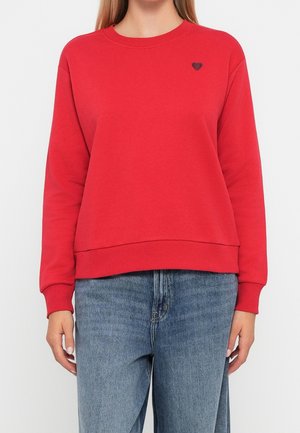 Czerwona bluza z długim rękawem i okrągłym dekoltem, z małą czarną haftowaną serduszkiem na lewej piersi, noszona z niebieskimi dżinsami.